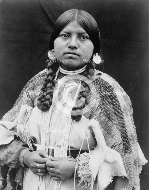 Cayuse woman, half-length portrait, standing, facing front, braids, shell disk earrings..., c1910. Creator: Edward Sheriff Curtis.