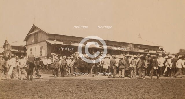 Crowds of people in front of buildings at the marketplace in the center of town..., (1899?). Creator: Eleanor Lord Pray.