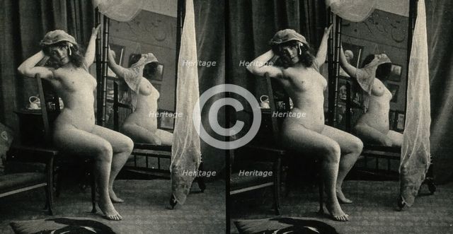 A woman posing naked in a photographic studio, sitting on a chair in front of a looking-glass, c1900 Creator: Unknown.