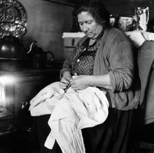 A woman sewing a shirt by the stove, London, 1953. Creator: Henry Grant