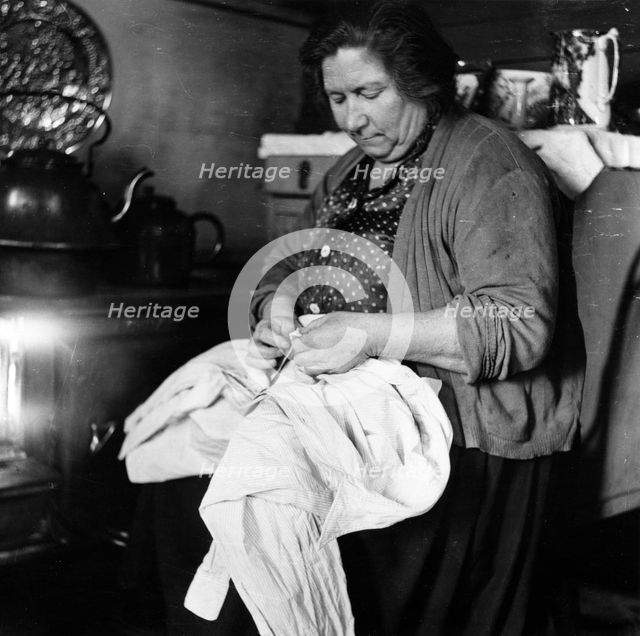 A woman sewing a shirt by the stove, London, 1953. Creator: Henry Grant.