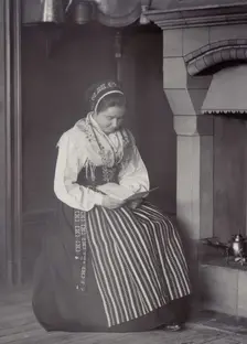 A woman sits in front of a fireplace and reads dressed in a Dalarna costume, 1886-1920. Creator: Helene Edlund