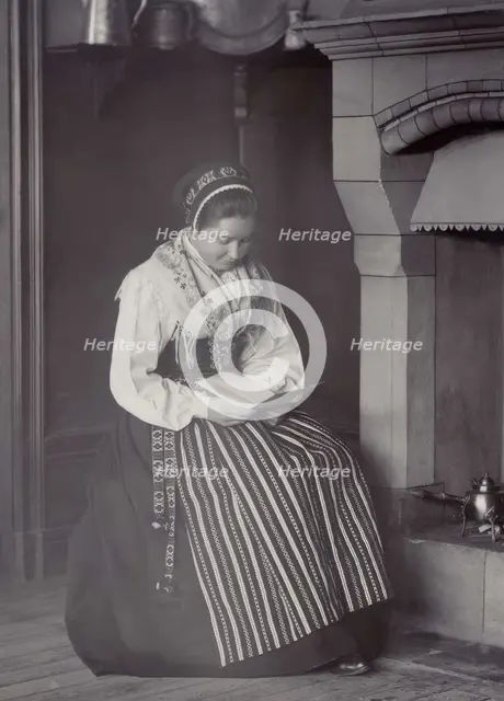 A woman sits in front of a fireplace and reads dressed in a Dalarna costume, 1886-1920. Creator: Helene Edlund.