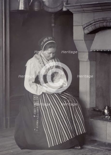 A woman sits in front of a fireplace and reads dressed in a Dalarna costume, 1886-1920. Creator: Helene Edlund.