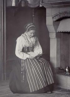 A woman sits in front of a fireplace and reads dressed in a Dalarna costume, 1886-1920. Creator: Helene Edlund