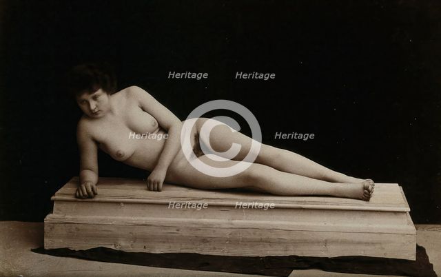 A young woman posing naked in a photographic studio, lying on a wooden plinth, approx 1900. Creator: Unknown.