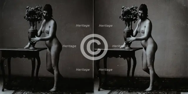 A young woman posing naked in a photographic studio, standing in front of a table on which..., c1900 Creator: Unknown.
