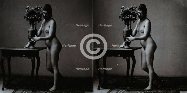 A young woman posing naked in a photographic studio, standing in front of a table on which..., c1900 Creator: Unknown.