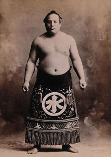 A young sumo wrestler posing in a photographic studio, wearing a richly decorated apron..., c1900. Creator: Unknown