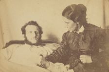 A bearded man lying in bed, his hand held by his wife who is seated next to the bed, 1870-79. Creator: Unknown