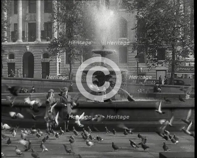 A Boy and a Girl Feeding Pigeons by One of the Fountains in Trafalgar Square with..., 1939. Creator: British Pathe Ltd.