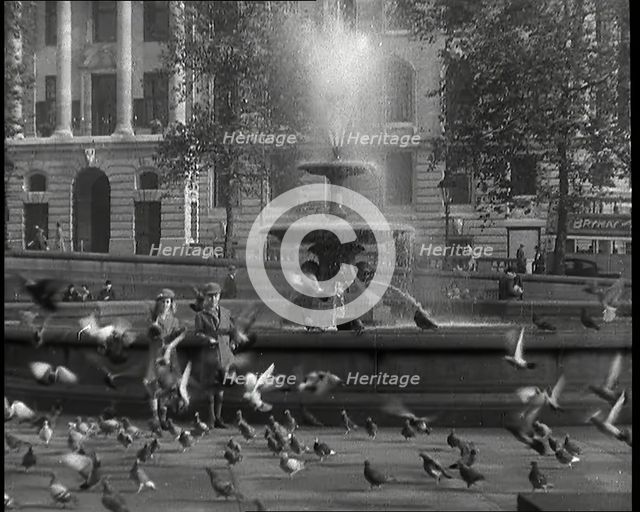 A Boy and a Girl Feeding Pigeons by One of the Fountains in Trafalgar Square with..., 1939. Creator: British Pathe Ltd.