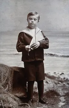 A boy with Down's syndrome, dressed in a sailor suit, standing against a nautical... Creator: Kent Coast Studio
