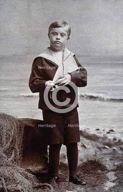 A boy with Down's syndrome, dressed in a sailor suit, standing against a nautical... Creator: Kent Coast Studio.