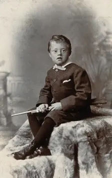 A boy with Down's syndrome, holding a rifle and sitting on a fur carpet, c1900. Creator: RW Morris