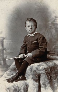 A boy with Down's syndrome, holding a rifle and sitting on a fur carpet, c1900. Creator: RW Morris