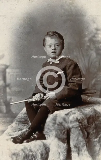 A boy with Down's syndrome, holding a rifle and sitting on a fur carpet, c1900. Creator: RW Morris.