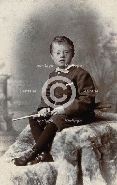 A boy with Down's syndrome, holding a rifle and sitting on a fur carpet, c1900. Creator: RW Morris.