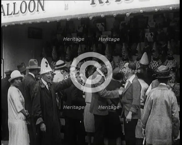 A Crowd of People Standing in Front of a Carnival Game. A Man in a Clown's Hat Encourages..., 1924. Creator: British Pathe Ltd.