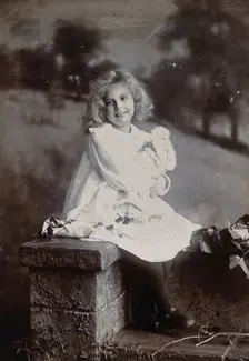 A girl, holding a doll, sitting on a wall, c1890s. Creator: Lock & Whitfield