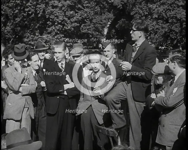 A Group of Male Civilians and One Soldier Listen to a Man With a Cigarette Standing on a..., 1938. Creator: British Pathe Ltd.