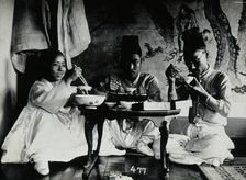 A group of people eating a meal, seated behind a small table, in front of a painted screen, c1900. Creator: Unknown