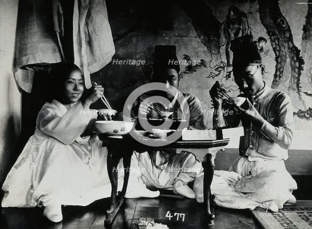 A group of people eating a meal, seated behind a small table, in front of a painted screen, c1900. Creator: Unknown.