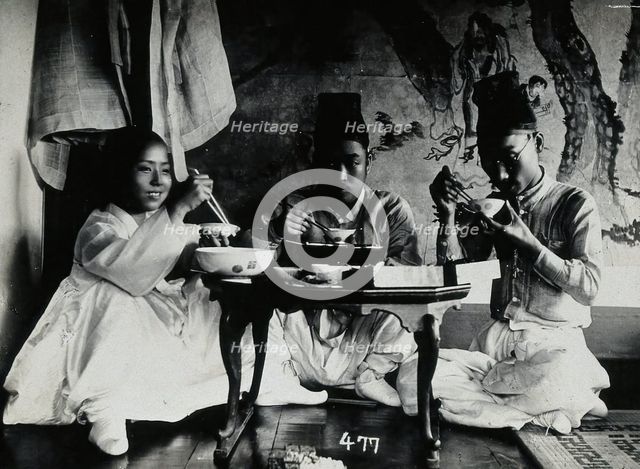 A group of people eating a meal, seated behind a small table, in front of a painted screen, c1900. Creator: Unknown.