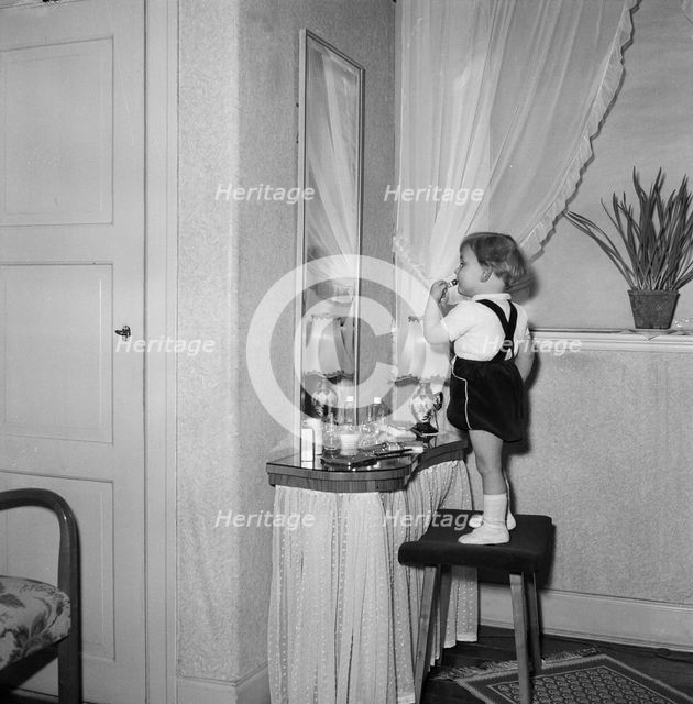 A little girl uses her mother's lipstick in front of a mirror, Sweden. Creator: Unknown.