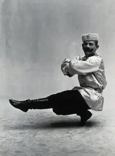 A man, in a satin tunic and fur hat, dancing a "Cossack" measure in a studio setting, 1899-1908. Creator: Unknown