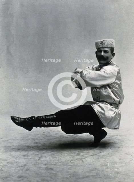 A man, in a satin tunic and fur hat, dancing a "Cossack" measure in a studio setting, 1899-1908. Creator: Unknown.