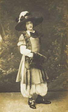 A man in drag wearing a very large hat with a veil, stands in front of a painted..., 1911. Creator: Unknown