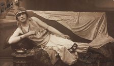 A man in drag poses wearing delicate attire; curled up on a piece of furniture, [1910-1919?]. Creator: Unknown