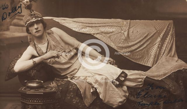 A man in drag poses wearing delicate attire; curled up on a piece of furniture, [1910-1919?]. Creator: Unknown.