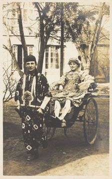 A man in ornate drag sits in a dog-cart and is pulled along the road by...(between 1910 and 1919?). Creator: Unknown