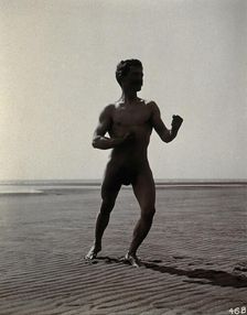 A man posing naked in a boxing stance with his fists raised, on a deserted beach, c1900. Creator: Unknown