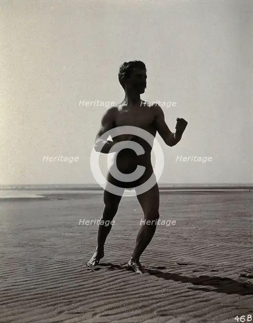A man posing naked in a boxing stance with his fists raised, on a deserted beach, c1900. Creator: Unknown.