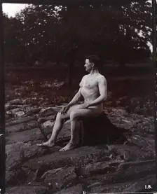 A man posing naked, seated on a rocky outcrop in a leafy landscape, c1900. Creator: Unknown