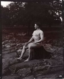 A man posing naked, seated on a rocky outcrop in a leafy landscape, c1900. Creator: Unknown