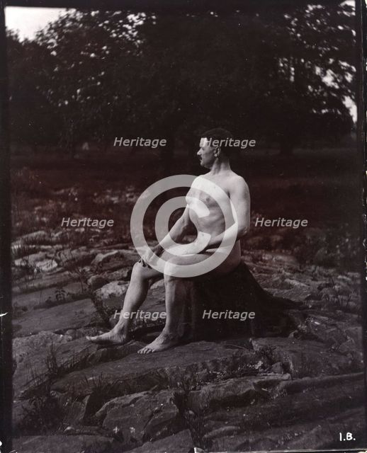 A man posing naked, seated on a rocky outcrop in a leafy landscape, c1900. Creator: Unknown.