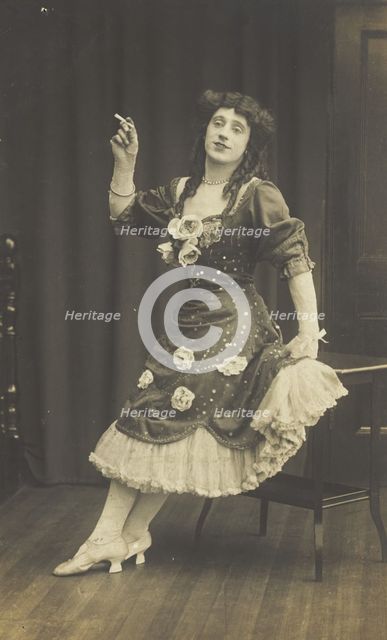 A performer in drag, wearing a large flowery dress, poses with a cigarette on stage, c1908-1910. Creator: Unknown.