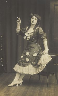 A performer in drag, wearing a large flowery dress, poses with a cigarette on stage, c1908-1910. Creator: Unknown