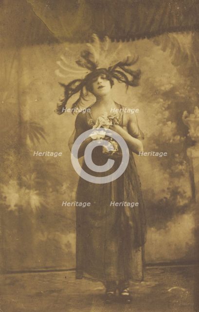 A performer in drag poses on stage wearing a head garment with sprawling feathers, [1910-1919?]. Creator: Unknown.