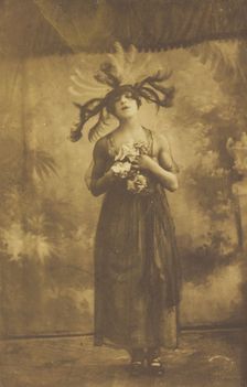 A performer in drag poses on stage wearing a head garment with sprawling feathers, [1910-1919?]. Creator: Unknown