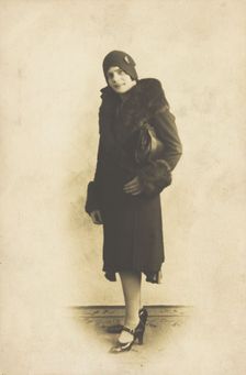 A person poses wearing a long coat with fur trim and a hat with a brooch, [between 1920 and 1929?]. Creator: Unknown