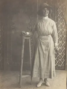 A sailor in drag leans on a high stool, in front of patterned scenery, (between 1910 and 1919?). Creator: Unknown