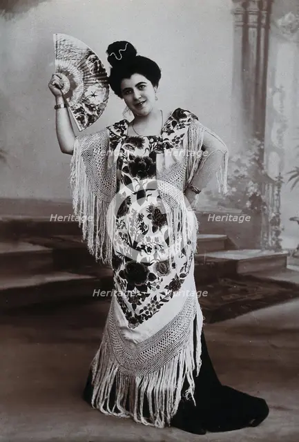 A standing woman, holding aloft a fan, wearing a fringed shawl, in front of a painted..., c1900. Creator: Unknown.