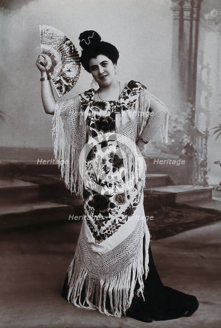 A standing woman, holding aloft a fan, wearing a fringed shawl, in front of a painted..., c1900. Creator: Unknown.