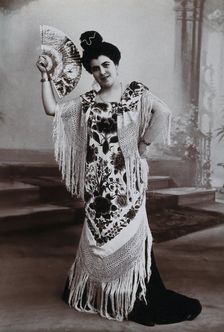 A standing woman, holding aloft a fan, wearing a fringed shawl, in front of a painted..., c1900. Creator: Unknown
