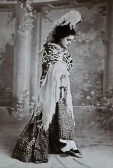 A standing woman, posed with an upraised fan and pointed toe, wearing a fringed shawl, in..., c1900. Creator: Unknown